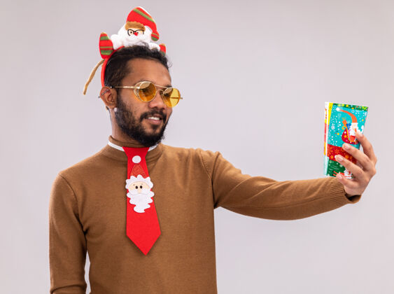 站着一个非洲裔美国人 穿着棕色毛衣 头戴圣诞老人圈 打着有趣的红色领带 手里拿着五颜六色的纸杯 面带微笑地看着它 站在白色的背景上搞笑美国人头