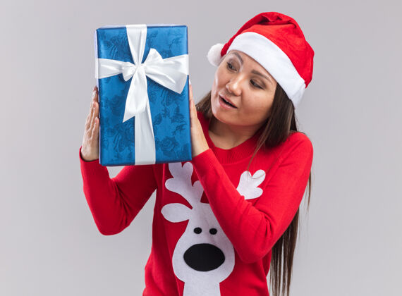 盒子印象深刻的年轻亚洲女孩戴着圣诞帽 拿着毛衣 看着白色背景上孤立的礼盒举行礼物毛衣