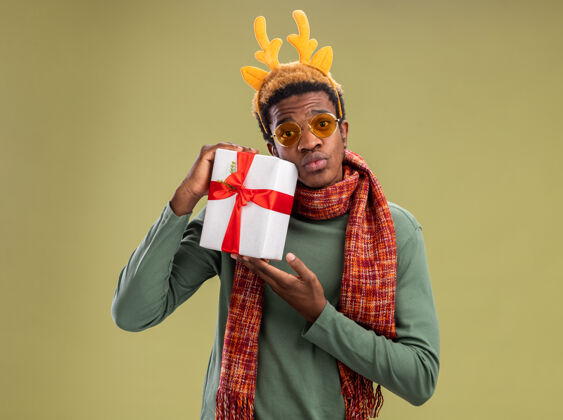 美国一个非洲裔美国人 戴着滑稽的边 脖子上围着鹿角和围巾 手里拿着圣诞礼物 困惑地站在绿色的背景上看着摄像机鹿搞笑角