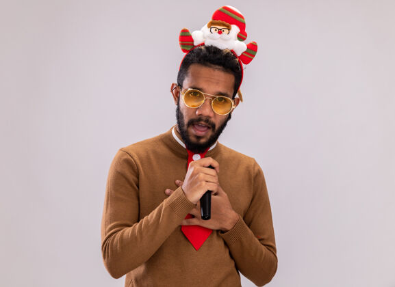 领带一个身穿棕色毛衣 头戴圣诞老人耳环 打着有趣的红色领带的非洲裔美国人拿着麦克风 站在白色背景上困惑地看着摄像机困惑圣诞老人美国人
