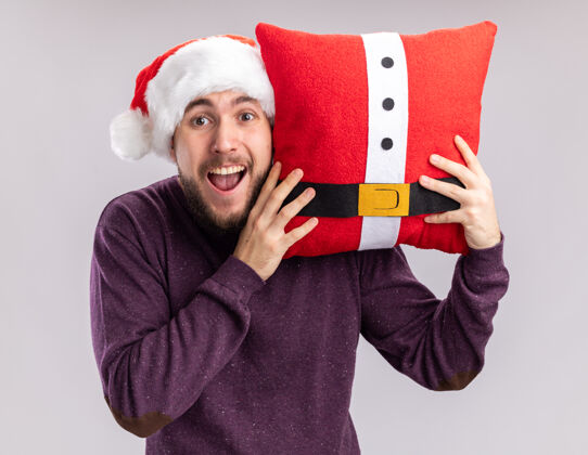 抱着快乐的年轻人穿着紫色毛衣 戴着圣诞帽 抱着滑稽的枕头 站在白色的背景下 惊讶地看着镜头惊讶年轻毛衣