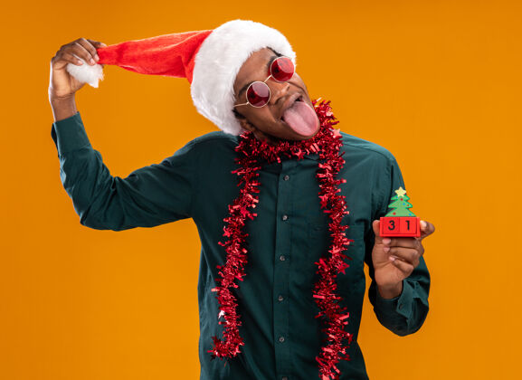 快乐快乐的非洲裔美国人戴着圣诞帽 戴着花环 戴着太阳镜 手里拿着新年日期的玩具方块 舌头伸出 站在橙色的背景上日期舌头帽子