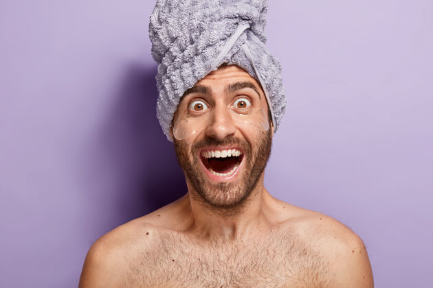 抗衰老眼睛下面垫着垫子的惊喜男人 赤膊上阵站在紫色背景下 头戴毛巾 关心脸部皮肤应用情绪健康