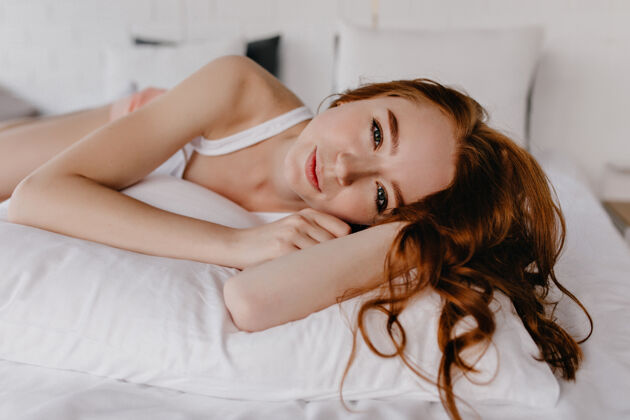 人浪漫的年轻女士 一头闪亮的姜黄色头发 在枕头上摆姿势身着白色睡衣的心醉神迷的女模特的室内照片房间室内快乐