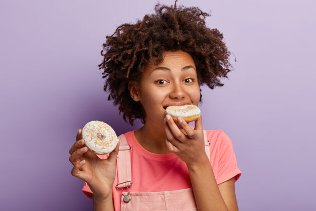 肖像饥肠辘辘的非洲女人咬着鲜亮的甜甜圈 有不健康的营养 无法想象没有甜点的生活 卷发 不节食 孤立在紫色的墙上非洲零食青少年