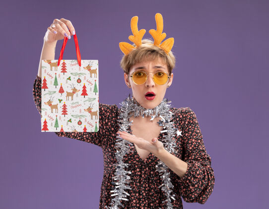 快乐关心的年轻漂亮女孩戴着驯鹿鹿角头带 脖子上戴着金箔花环 戴着眼镜 拿着并指着圣诞礼品袋 看着隔离在紫色背景上的相机圣诞节周围头带