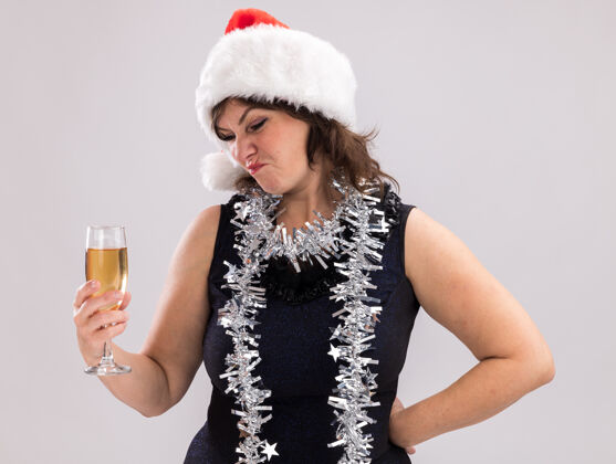帽子皱眉的中年妇女戴着圣诞帽 脖子上戴着金箔花环 手放在腰上 手放在白色背景上 手拿着香槟 看着杯子玻璃杯香槟保持