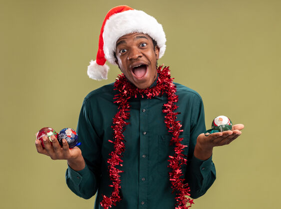 帽子戴着圣诞帽 戴着花环 拿着圣诞球的非裔美国人站在绿色背景下 面带微笑地看着摄像机站着圣诞节圣诞老人