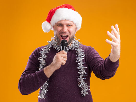帽子中年男子戴着圣诞帽 脖子上戴着金属丝花环 拿着麦克风 看着摄像机 手举着手 在橙色背景上孤立地唱歌保持中年周围
