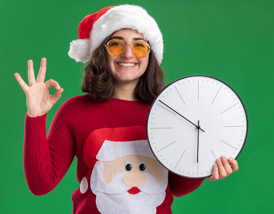 举行穿着圣诞毛衣的小女孩戴着圣诞帽戴着眼镜拿着挂钟看着相机脸上挂着微笑的ok标志站在绿色的背景上站着女孩年轻