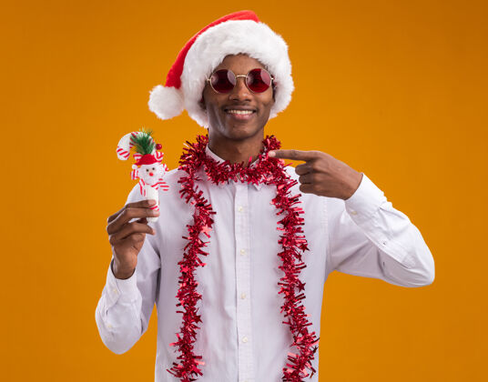 金属丝微笑着的年轻的非洲裔美国人戴着圣诞帽 戴着一副眼镜 脖子上戴着金箔花环 拿着并指着糖果手杖饰品 看着孤立在橙色背景上的相机指着圣诞老人微笑