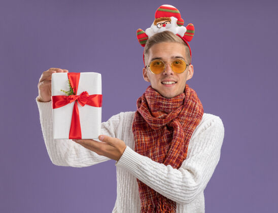 头带高兴的年轻帅哥戴着圣诞老人的头带和围巾拿着礼包看着相机孤立的紫色背景圣诞老人包抱