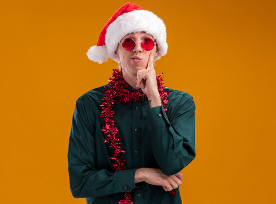 眼镜体贴的年轻金发男子戴着圣诞帽 戴着眼镜 脖子上戴着金属丝花环 看着相机 手放在下巴上 橙色背景上 留有复印空间圣诞老人保持年轻