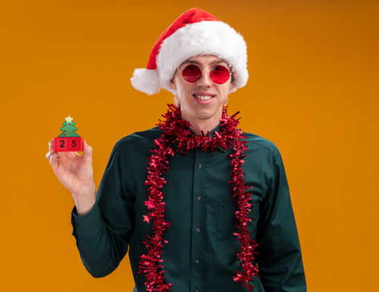 圣诞老人微笑着的金发年轻人戴着圣诞帽 戴着眼镜 脖子上戴着金属丝花环 手里拿着圣诞树玩具 手里拿着日期 看着隔离在橙色背景上的相机周围微笑花环
