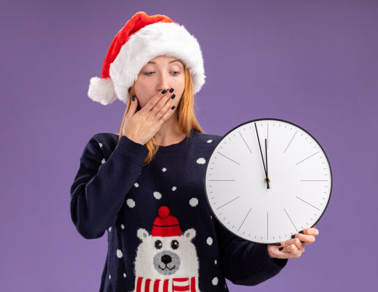 圣诞节吓得年轻漂亮的女孩穿着圣诞毛衣 戴着圣诞帽 拿着挂钟看 嘴巴上用手隔离着紫色的背景害怕年轻毛衣