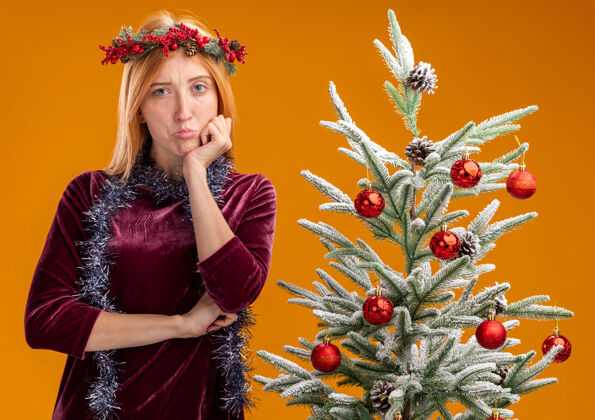 花环站在圣诞树旁的悲伤的年轻漂亮的女孩穿着红色的裙子 脖子上戴着花环 手放在下巴上 橙色的背景上孤立着下巴圣诞节悲伤