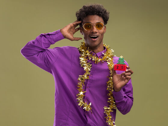 圣诞节令人印象深刻的年轻非裔美国人戴着眼镜 脖子上戴着金属丝花环 手里拿着圣诞树玩具 手里拿着日期 看着相机 手放在头上 在橄榄绿的背景下与世隔绝树保持举行