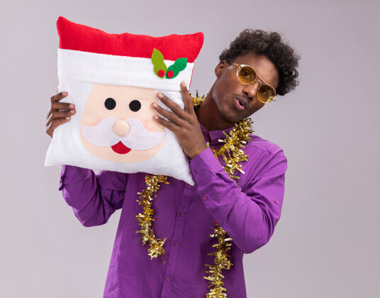 非裔美国人令人印象深刻的美国黑人年轻人戴着眼镜 脖子上戴着金箔花环 抱着圣诞老人的枕头 看着隔离在白色背景上的相机花环年轻的金属丝