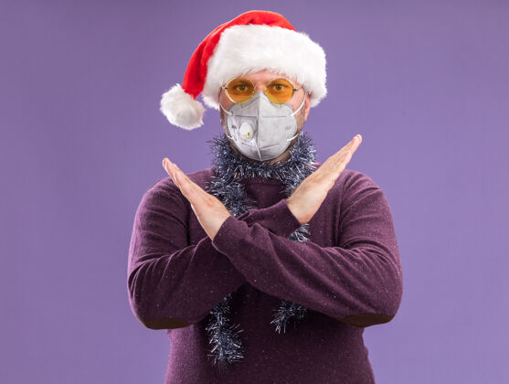 周围严肃的中年男子戴着圣诞帽 戴着防护面具 脖子上戴着金属丝花环 戴着眼镜 看着镜头 做着没有动作的紫色背景隔离面具做保护