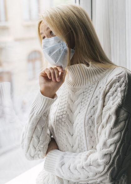 大流行大流行期间戴着医用口罩在家的女人透过窗户看Covid19妇女流行病