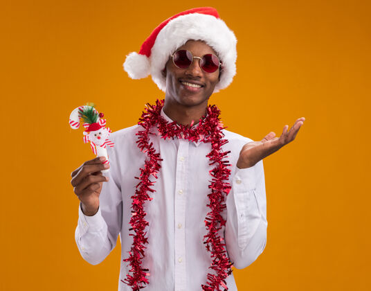 糖果微笑着的年轻的非洲裔美国人戴着圣诞帽 戴着眼镜 脖子上戴着金箔花环 手里拿着糖果手杖 看着相机 在橙色的背景上显示出一只孤立的空手手杖装饰品圣诞老人