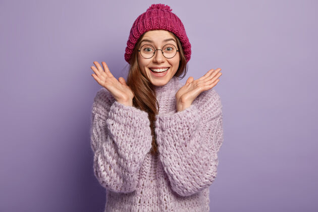 穿着照片中幸福感性的欧洲女人双手比划 掌心相扣 戴着圆眼镜 戴着冬日礼帽 针织套头衫 隔着紫色的墙好情绪概念表情季节姿势