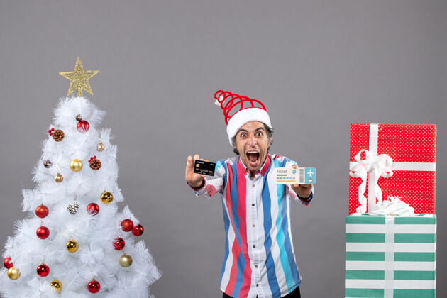 抱着前视图：手持卡片和旅行票的男子在圣诞树周围大声喊道 并赠送礼物旅行树票