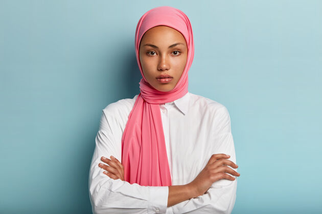 宗教半身镜头的严肃自信的女人戴着传统的粉色头巾 双臂交叉 穿着白衬衫 站在蓝色的墙上 饶有兴趣地聆听对话者宗教阿拉伯语女人头像