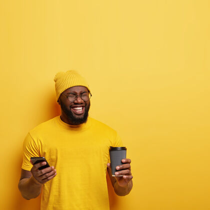 交谈照片中的快乐快乐黑皮肤的家伙从快乐中闭上眼睛 戴着黄色时髦的帽子和t恤 在社交网络上留言 拿着现代手机和咖啡走 对一些有趣的东西咯咯笑自由空间男朋友设备
