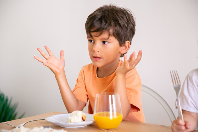 叉子可爱的小男孩吃着生日蛋糕 喝着橙汁可爱的孩子坐在饭厅的桌子旁 举手看着别处童年 庆祝和节日的概念蛋糕肖像庆祝