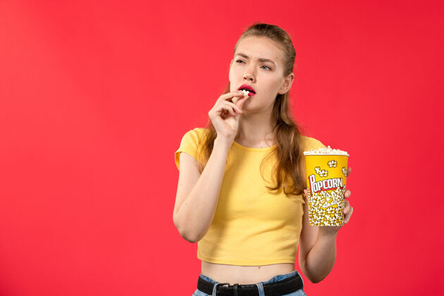 观点正面图年轻女性在电影院拿着爆米花包吃着淡红墙电影院里的女性趣味电影漂亮表演者浅红色