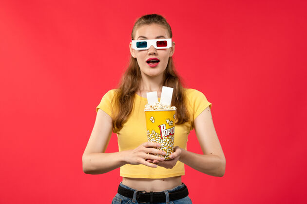 女性正面图年轻女子在电影院拿着爆米花包 戴着d型太阳镜 拿着红桌电影票 电影院里的电影女孩电影票电影