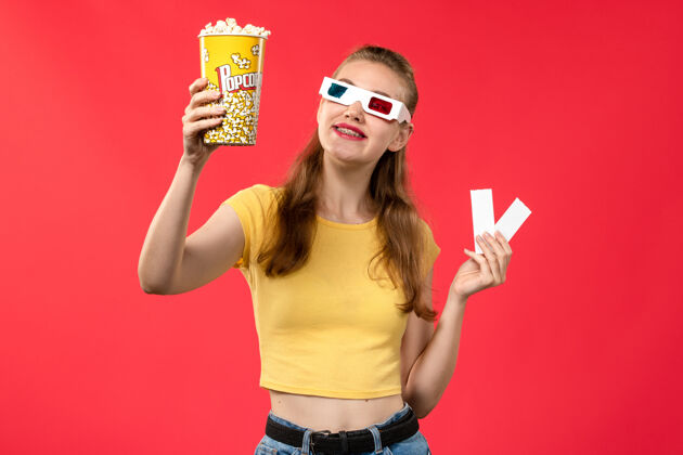 爆米花正面图年轻女性在电影院拿着爆米花和票戴着d太阳镜在红墙电影院看电影女性电影院剧院