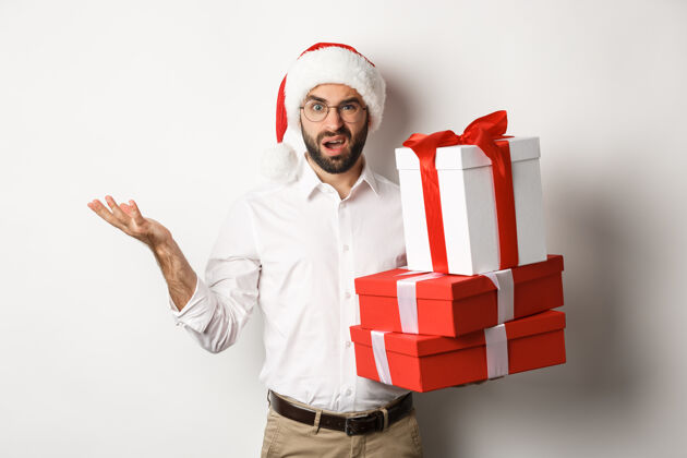情绪化圣诞快乐 节日概念男人拿着圣诞礼物 困惑地耸耸肩 戴着圣诞帽站在白色背景下成人庆祝兴奋