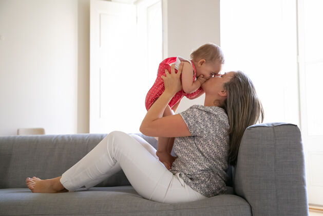 幼儿慈爱的母亲坐在沙发上 用爱亲吻她的小女儿快乐的女婴用手掌合上脸长发的母亲用双手抱着婴儿家庭和母亲的概念年轻孩子妈妈