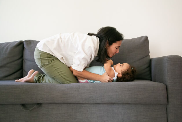 视图快乐的黑发妈妈抱着可爱的小女儿在灰色沙发上侧视图父母和童年的概念年轻室内童年