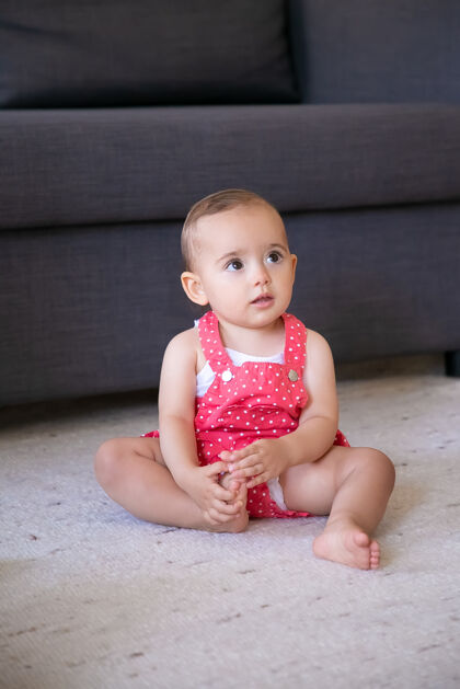 沉思可爱的小宝宝赤脚坐在客厅的地毯上可爱体贴的女孩穿着红色的工作服短裤看着别人 抚摸着脚周末 童年和在家的概念天真情感新生儿