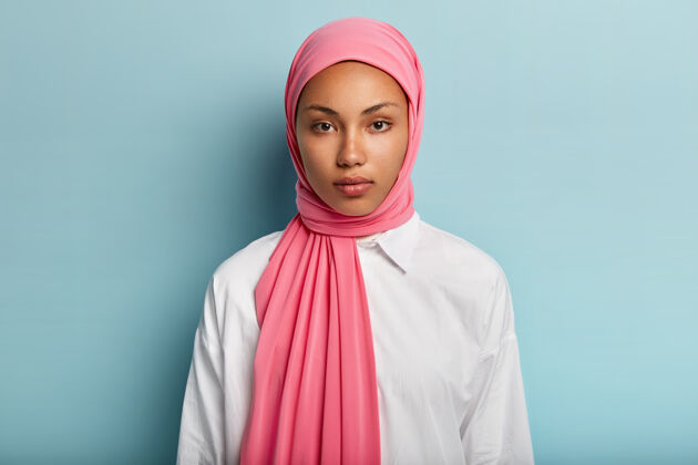 肖像表情冷静严肃的阿拉伯女人 皮肤黝黑 裹着粉色面纱 穿着白色衬衫 没有化妆 自然美丽 蓝墙之上的模特女士特写镜头夹克凝视风格