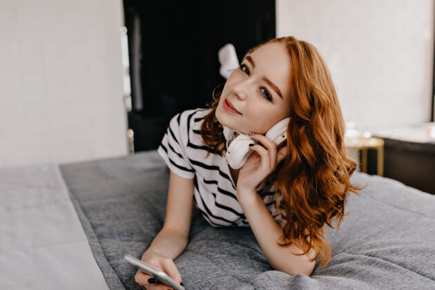 放松幸福的红发女孩躺在床上拿着电话有兴趣的高加索女人拿着白色耳机成人卧室积极