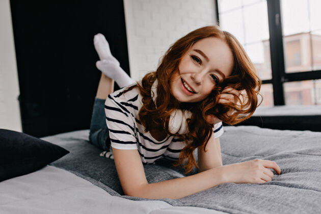 房间美丽的年轻女模特躺在床上优雅的卷发女孩摆出可爱的微笑女士耳机红发