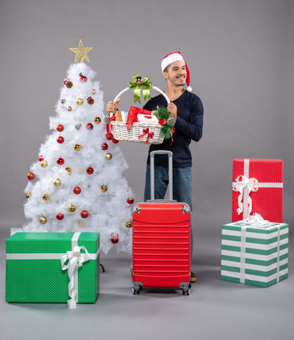 购物者微笑的年轻人戴着圣诞帽 双手捧着礼品篮 靠近灰色的圣诞树礼物包微笑的年轻人