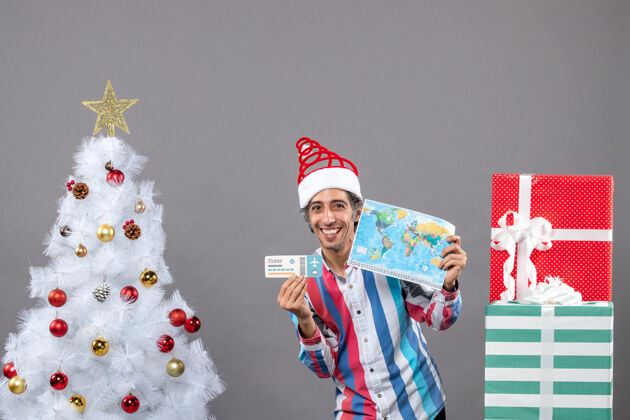 圣诞树正面图微笑的戴着圣诞帽的男人拿着世界地图和旅行票假期有趣男