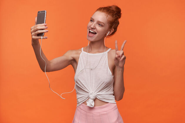 发型迷人的年轻readhead女性 发髻 头戴耳机 举手表示和平 一边愉快地看着相机 一边用橙色背景的智能手机自拍音乐头发智能手机