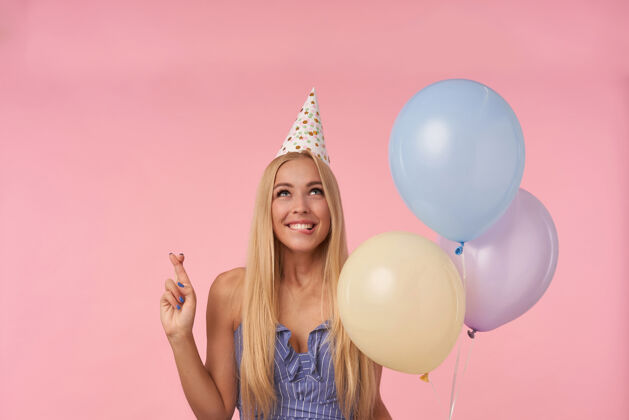 正面快乐迷人的年轻金发女性许下愿望 交叉手指祝好运 手持一束氦气球 穿着蓝色夏装和生日帽在粉色背景上摆姿势头发迷人条纹