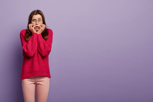 害怕紧张紧张的女人横拍感觉神经质 咬指甲 穿超大的红色长款套头衫 对惊人的消息做出反应 站在紫色的墙壁空白处获取信息编织考试神经质