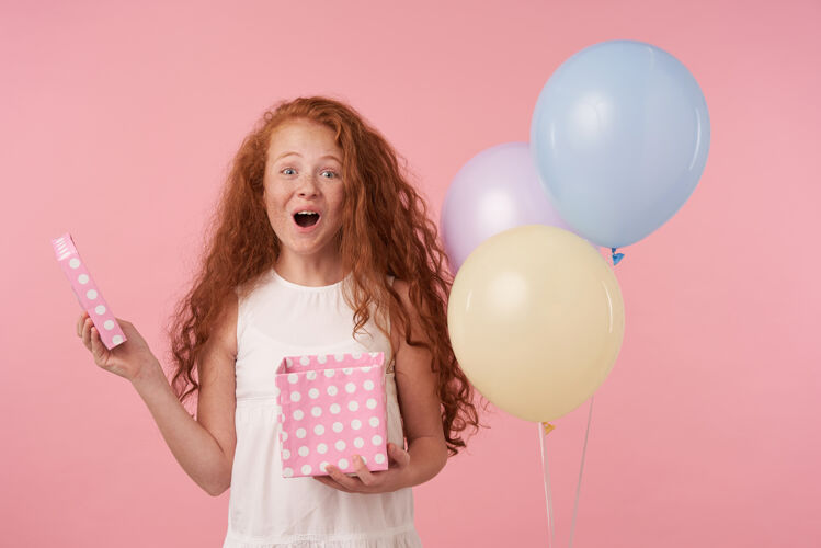 节日快乐的红头发女孩的肖像 长卷发 穿着优雅的衣服 手里拿着礼物盒 兴奋地打开它 快乐地看着粉红色背景下的相机卷曲孩子孩子