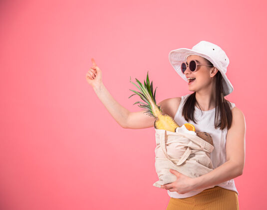 太阳镜戴着白色帽子和太阳镜的时髦女孩微笑着 手里拿着一个装有异国水果的环保袋模型女人异域风情
