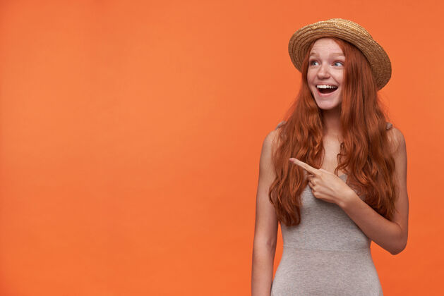 女士摄影棚拍摄了一个快乐的年轻红发女子 留着一头长发 穿着灰色衬衫 戴着草帽 在橙色背景上摆姿势 举起食指 看着一边 脸上露出惊讶的表情张开漂亮长