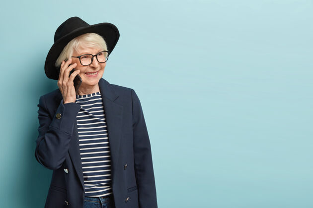 女士美丽的白发女士在工作日后有电话交谈 通过申请打电话 讨论新的成功完成的任务 戴着时髦的头饰 隔着蓝色的墙谈话高兴成熟