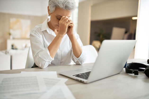 头部一张室内照片 一位沮丧 不快乐的中年女商人坐在办公桌前 手放在脸上 低头看报纸衰老技术桌子